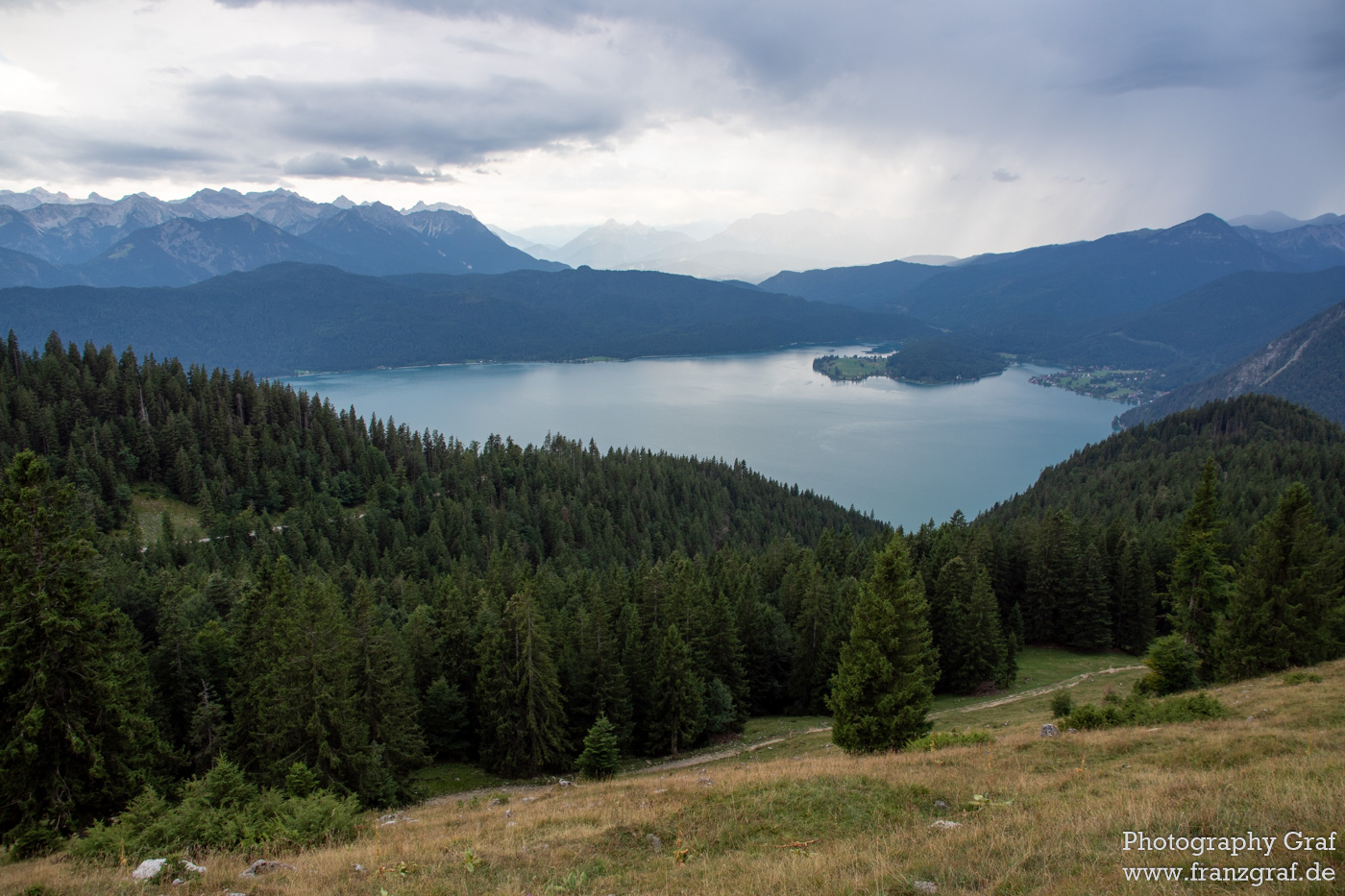 This breathtaking landscape is comprised of a crystal clear lake surrounded by tall, lush trees and a mountainous backdrop that completes the perfect outdoor scene. The sky is a mix of light grey and white clouds, adding to the beauty of the view. The trees are a mix of green and brown, with some having leaves that are still vibrant and full of life. The lake has a tranquil atmosphere, adding a peaceful atmosphere to the area. The mountains are covered in snow, and the tops of some can be seen in the distance, creating a beautiful contrast with the green trees and blue sky. The scene is one of a kind and is sure to be remembered for years to come.