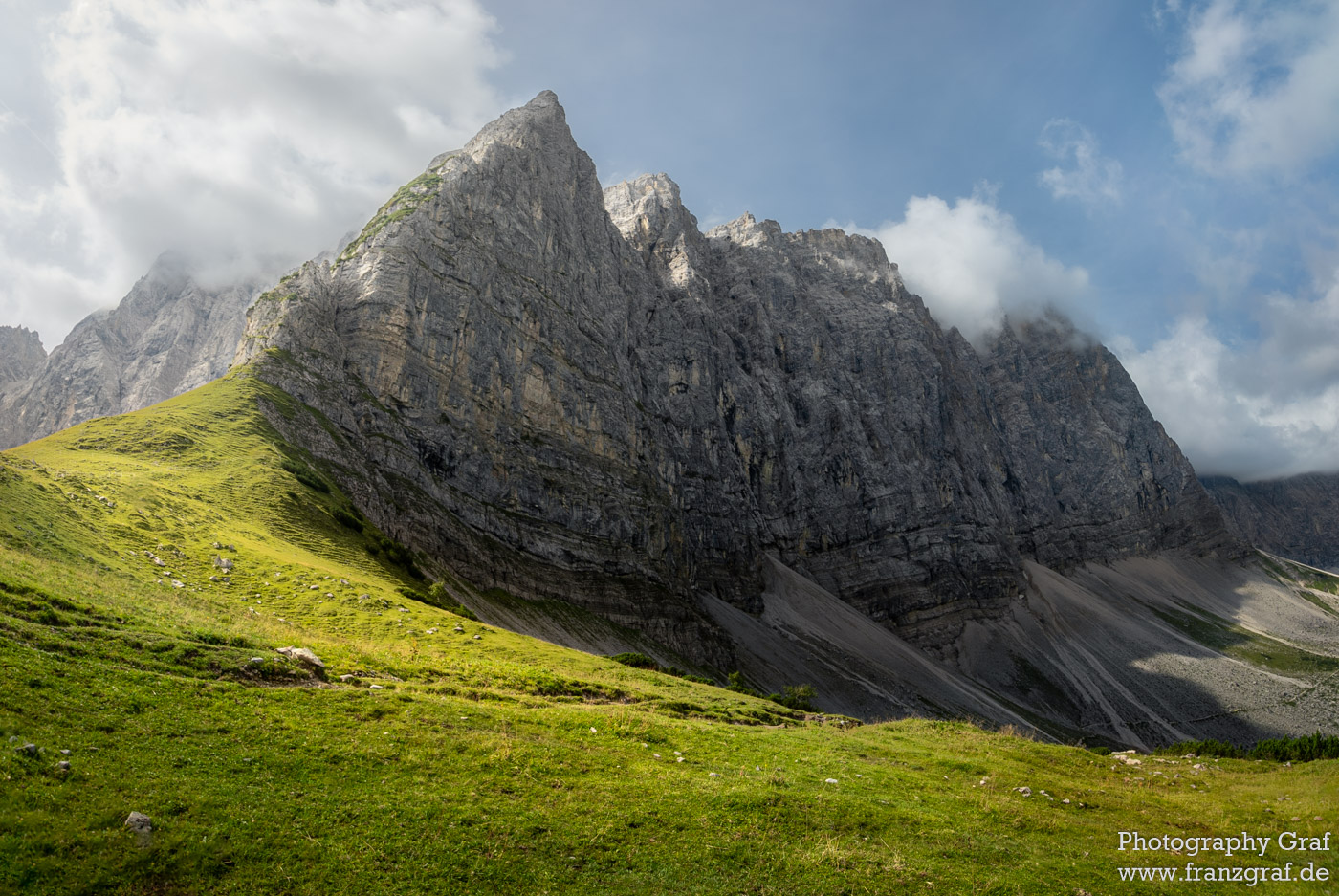 This picture shows a stunning outdoor mountain landscape with a mountain range stretching out in the background. The sky is filled with white, fluffy clouds, and the sun casts a bright light on a grassy hill at the forefront. The hill is dotted with clusters of rocks, giving the landscape a rugged texture. In the middle ground, a large rocky cliff creates a dramatic contrast against the bright green grass. The land gradually slopes down into a lush valley beyond the cliff. In the foreground, a person stands atop a dirt hill with a shadow stretching out behind them, providing a sense of scale and perspective. All of these elements combine to create an awe-inspiring scene that is sure to take your breath away.
