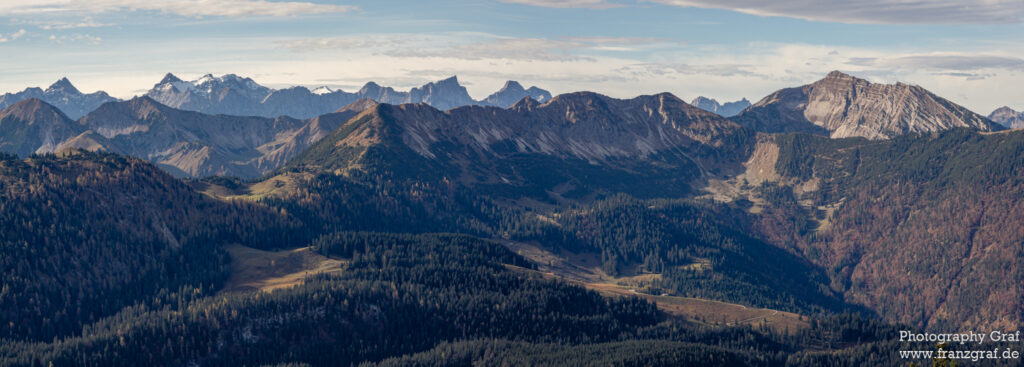 This is an enchanting panoramic view of a mountain range. The mountains are majestic and boast imposing peaks that pierce through the sky. The terrain is rugged, with an array of trees dotted across the landscape, adding a touch of green to the scene. The sky is vast and open, with fluffy clouds hovering above the mountain range, casting a soft glow over the scene. The atmosphere evokes a sense of tranquility and awe, making the picture a perfect example of the serene beauty of the outdoors. The image could be from a national park, given the untouched wilderness depicted. The view could also be from the Alps, considering the highland terrain and the snow on the mountaintops. The photograph excellently captures the raw beauty of nature, making one feel as if they are standing on a summit, taking in the breath-taking view.