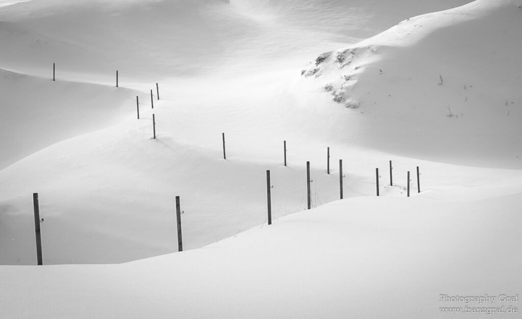 This stunning image captures a serene winter landscape blanketed in snow. The dominant feature of the photograph is a fence, almost lost amidst the white expanse, offering a stark contrast to the soft, snowy surroundings. The fence seems to be on a slope, suggesting the location could be a ski slope or a mountain. Nature's beauty is on full display, with the snow-covered setting exuding a sense of tranquility. The photograph is in black and white, enhancing the ethereal and foggy atmosphere. The absence of any people or animals adds to the sense of solitude and calmness. The scene is outdoors, and the weather seems to be freezing, suggesting it was taken in the heart of winter.