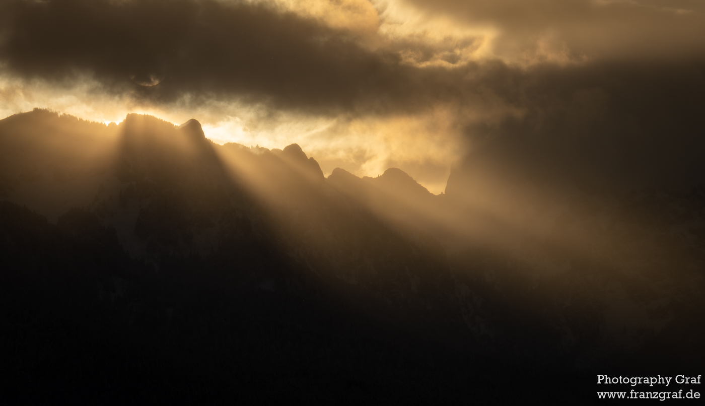 The image showcases a mountain range under a dramatic sky. Dark clouds loom, yet sunbeams break through, casting light on the misty mountainsides. The scene is a striking contrast of light and shadow, highlighting nature's raw beauty. The sun's rays create an otherworldly glow, emphasizing the rugged terrain. The photograph is a powerful depiction of nature's contrasts, evoking awe and wonder in its viewers. It's a visual narrative of resilience, where light overcomes darkness, and the mountains stand as enduring symbols of strength. The watermark credits the artist's ability to capture such a moment of natural splendor.