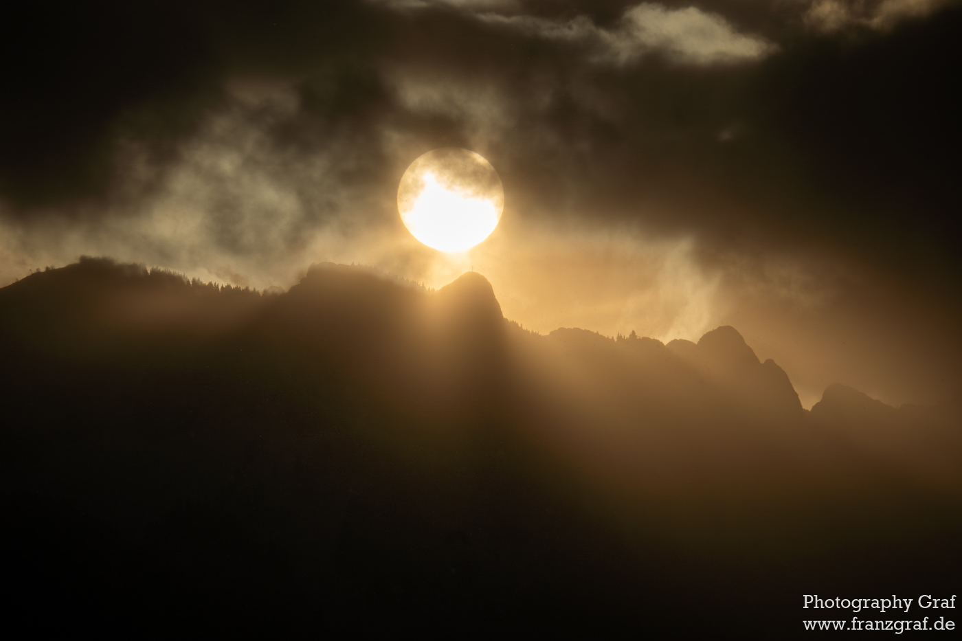 The image unfolds a dramatic natural tableau, where the sun's descent (or ascent) casts a radiant glow behind a mountain range. Dark clouds traverse the sky, yet the sun's brilliance pierces through, spotlighting the rugged peaks. This interplay of light and shadow, accentuated by the rays" creates a dynamic and almost three-dimensional effect. The mountains, silhouetted against the bright backdrop, stand as timeless witnesses to the spectacle. The scene is a powerful display of nature's contrasts, evoking awe and a sense of tranquility in its viewers. It's a visual poem, where each element contributes to the overall ethereal atmosphere, reminding us of the enduring beauty of the natural world. The watermark indicates the artist's skill in freezing this moment of awe-inspiring interplay between light and darkness.