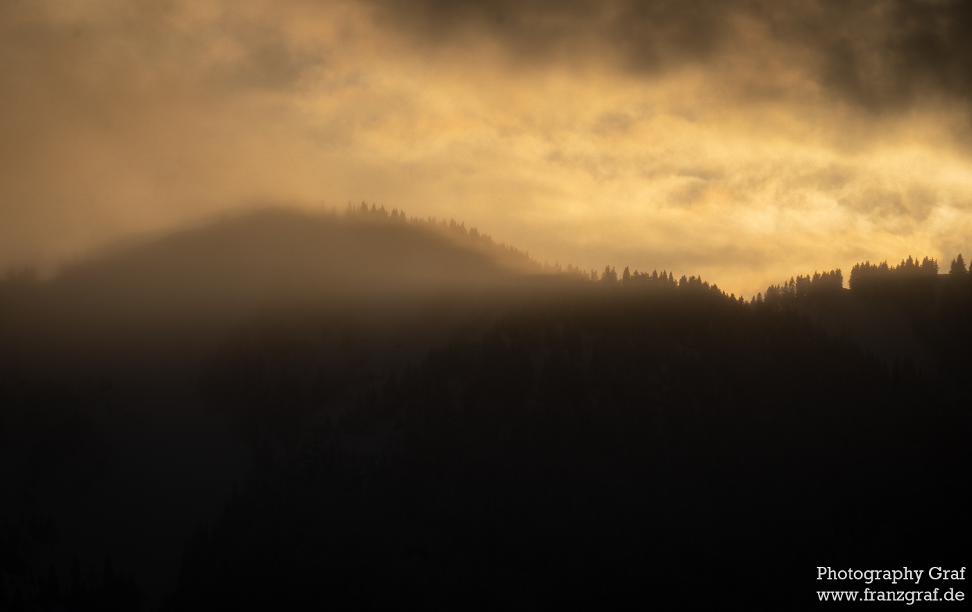 The image is a mesmerizing blend of nature's tranquility and drama. Mist shrouds forested mountains, while a golden sky suggests the quiet glow of dawn or dusk. The mountains' silhouettes peek through the mist, adding a sense of depth and mystery. Clouds weave dynamically through the scene, enhancing the landscape's ethereal quality. The photograph captures the dual essence of beauty and enigma in the natural world, inviting contemplation and wonder. It's a snapshot that beautifully conveys the silent dance between light and shadow, and the enduring allure of the wilderness. The watermark indicates the artist's skill in encapsulating such a powerful display of nature's contrasts.