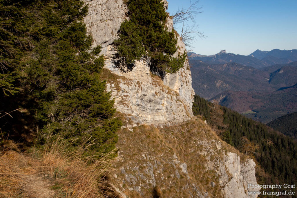 The photo offers a scenic view of a mountainous landscape, featuring a rocky cliff adorned with green trees in the foreground. A trail at the base of the cliff suggests a path for hiking or walking. The rugged textures of the rock face, along with the green coniferous trees, contrast beautifully against the multiple blue-shaded mountain ranges in the background. The clear sky above indicates fair weather, perfect for enjoying the outdoors.  It's a captivating scene that invites viewers to explore and appreciate the grandeur of nature. 