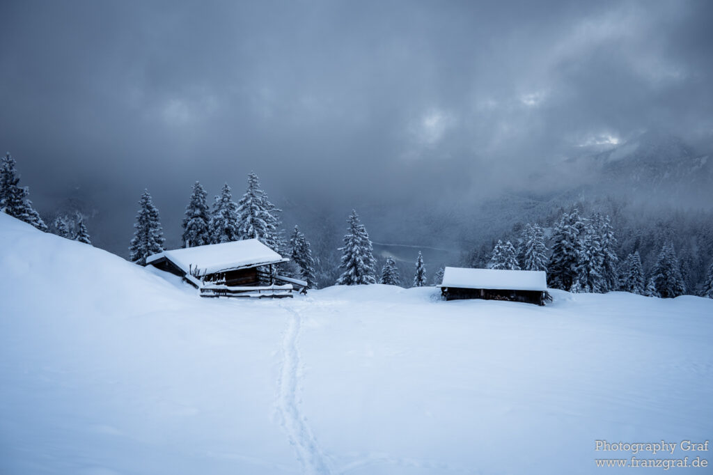 This image captures the serene beauty of a winter landscape. Blanketed in a thick layer of snow, the scene displays nature in its most peaceful form. In the foreground, we see a quaint cabin, its roof heavily laden with a fresh dusting of snow. The cabin is nestled amidst a dense gathering of trees, their branches sagging under the weight of the snow. In the distance, the snow-covered slopes of a mountain range add depth to the landscape. The sky above is a stark contrast to the white below, with its cloud-filled expanse hinting at the possibility of an impending winter storm. This image is a perfect blend of the raw power of nature and the tranquility that winter brings.