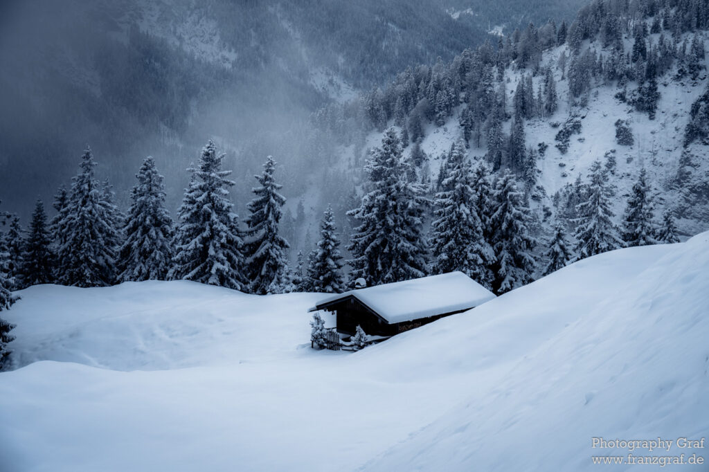 This beautiful image captures a serene winter scene viewed outdoors. The main focus is a cozy cabin nestled in an expanse of pristine, untouched snow. It's a freezing winter day, evident from the snowy landscape that surrounds the cabin. The snow seems to have recently fallen, as indicated by the glacial landform and the blizzard-like conditions. The distant trees and the slopes surrounding the cabin suggest a mountainous terrain, possibly a ski resort. There's also an obscured land vehicle near the cabin. The overall scene evokes a sense of peaceful isolation, a perfect escape for those who love nature and winter sports.