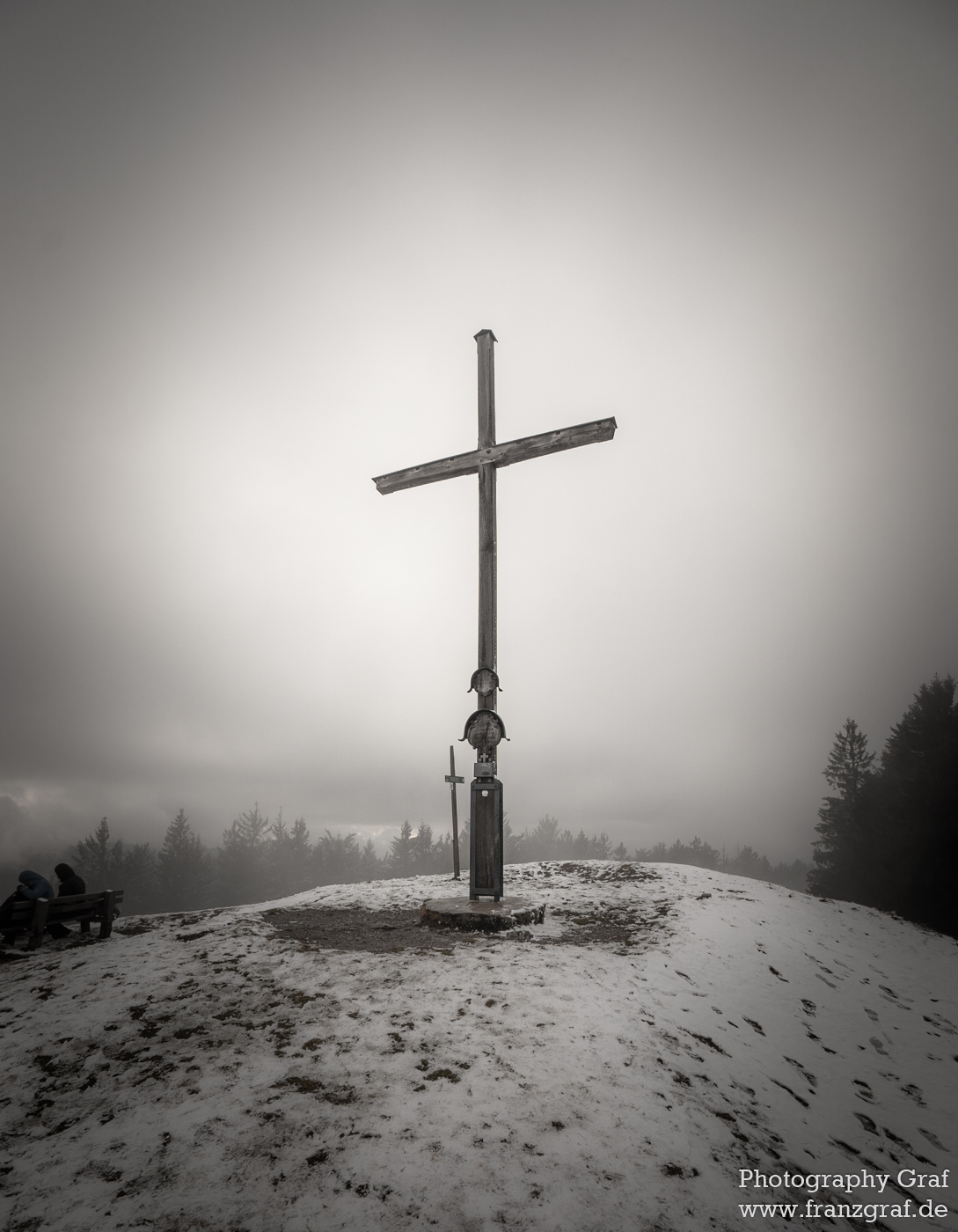 This image captures an evocative and serene winter scene, dominated by a large, solitary cross standing tall on a snowy hill. The sky overhead is vast and seems to blend seamlessly into the foggy landscape below, creating a hazy, dream-like atmosphere. The scene is devoid of vibrant colors, with varying shades of grey and white providing a monochromatic effect that further enhances the sense of solitude and tranquility. The ground beneath is blanketed in snow, dotted occasionally by gravestones, hinting that the cross might be a part of a cemetery. The image also features a street light, adding a slightly modern touch to the otherwise timeless setting. The overall mood of the picture is contemplative and somber, as if inviting viewers to pause and reflect.