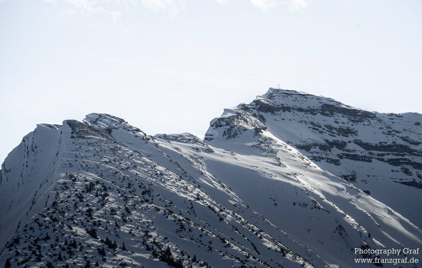 This image captures the raw beauty of nature in its most serene form. It features a majestic snow-covered mountain with a clear, radiant blue sky as the backdrop. The mountain's summit, slopes, and ridges are beautifully blanketed with a pristine layer of snow, making the glacial landform even more impressive. The terrain is also dotted with trees, adding a touch of life to the icy, white landscape. In the distance, one can see the outline of a mountain range, possibly the Alps, further enhancing the overall vista. This winter landscape is breathtaking, a perfect illustration of a peaceful, snowy day in the mountains.